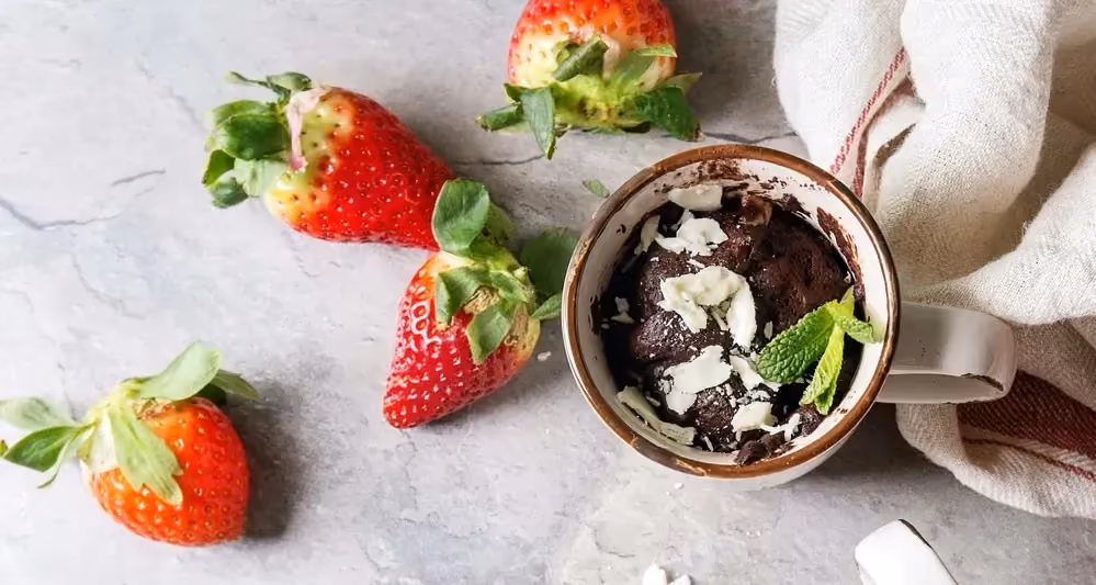 Un mugcake au chocolat est déposé sur un plan de travail. Il est accompagné de quatre grosses fraises. Une serviette blanche est présente sur le côté.