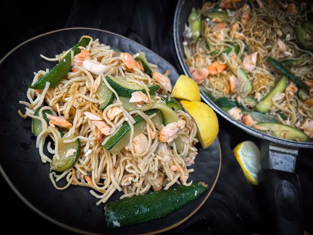 Dans une assiette se trouve les nouilles chinoises avec le saumon émietté et les courgettes coupées en fines lamelles. Du citron jaune est présent.