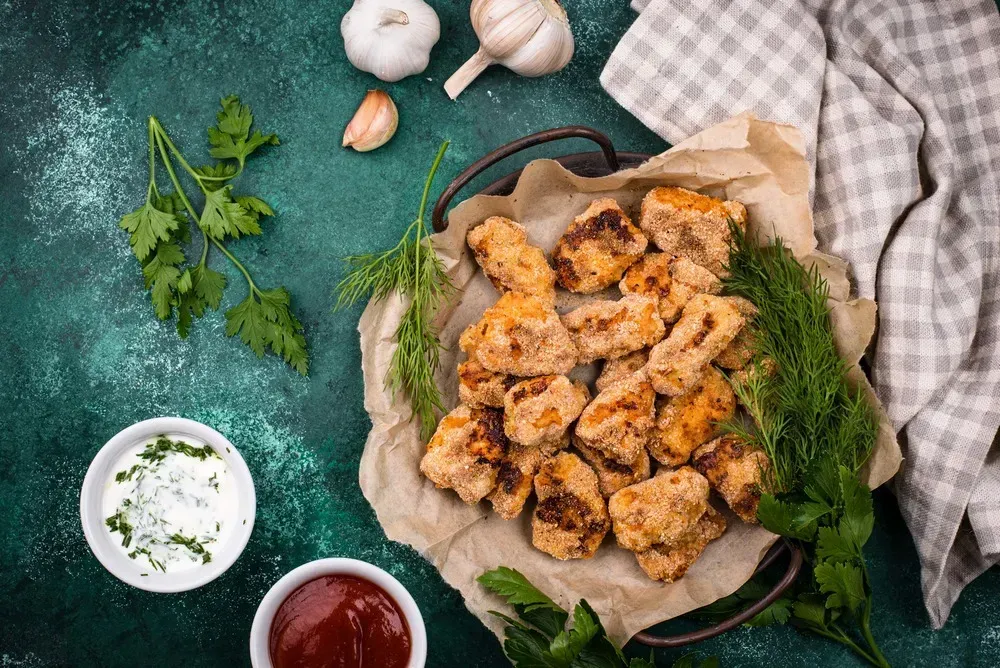 Small, nicely browned chicken nuggets are shown in the baking dish. Placed beside it are garlic and a sprig of parsley.