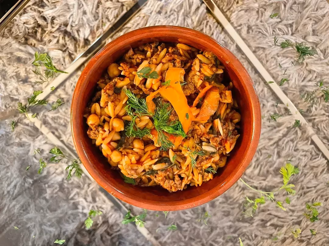 Le one pot orzo, épinards, boeuf et pois chiches est présenté dans un bol en céramique marron sur une table transparente.