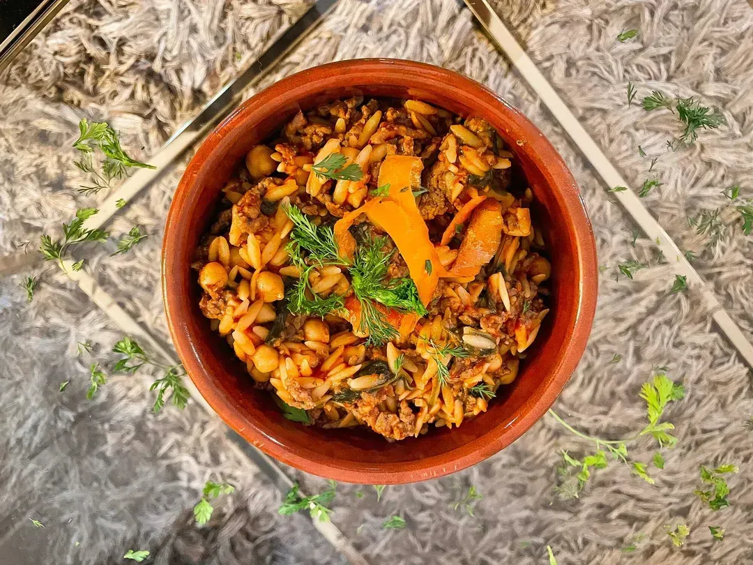 The one-pot orzo with spinach, beef, and chickpeas is presented in a brown ceramic bowl on a transparent table.