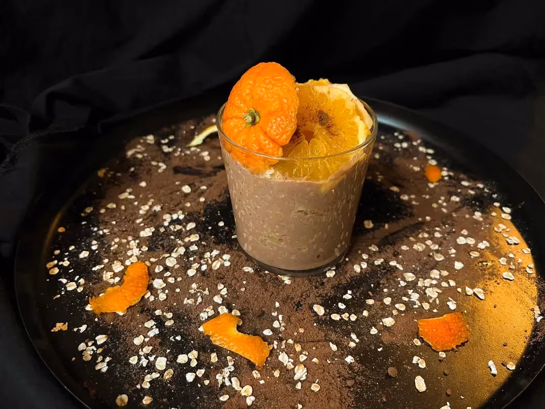 Sur un plat peu profond se trouve une verrine remplie par un overnight avec des flocons d'avoine, de l'orange, du chocolat et de la cannelle.