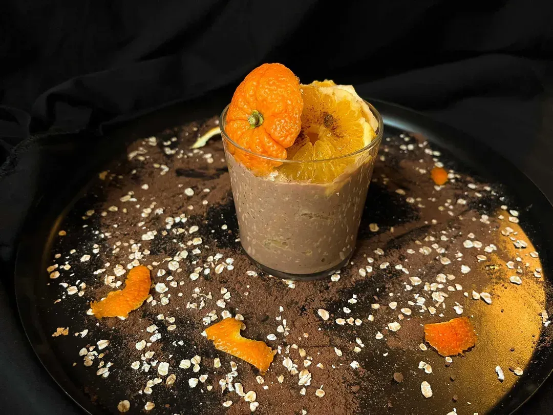 On a shallow plate sits a glass filled with overnight oats made with rolled oats, orange, chocolate, and cinnamon.