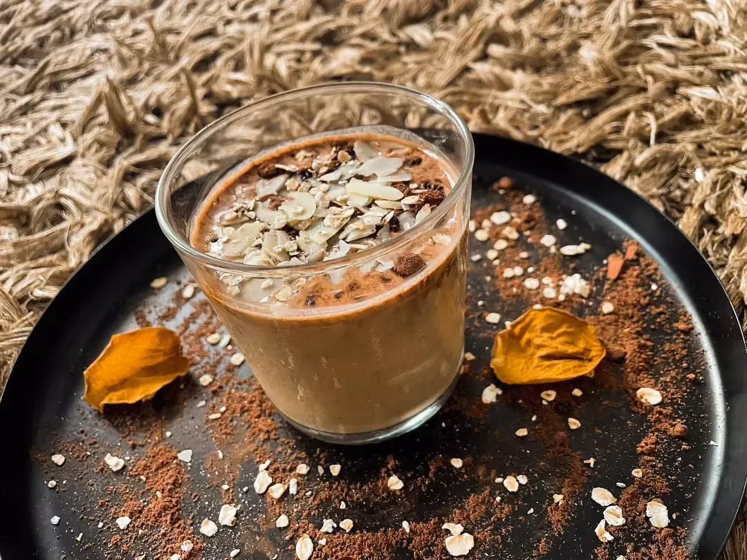 Une verrine posée sur un plat rond peu profond reçoit l'overnight composé d'avoine, café et chocolat. le topping est composé d'amandes effilées.
