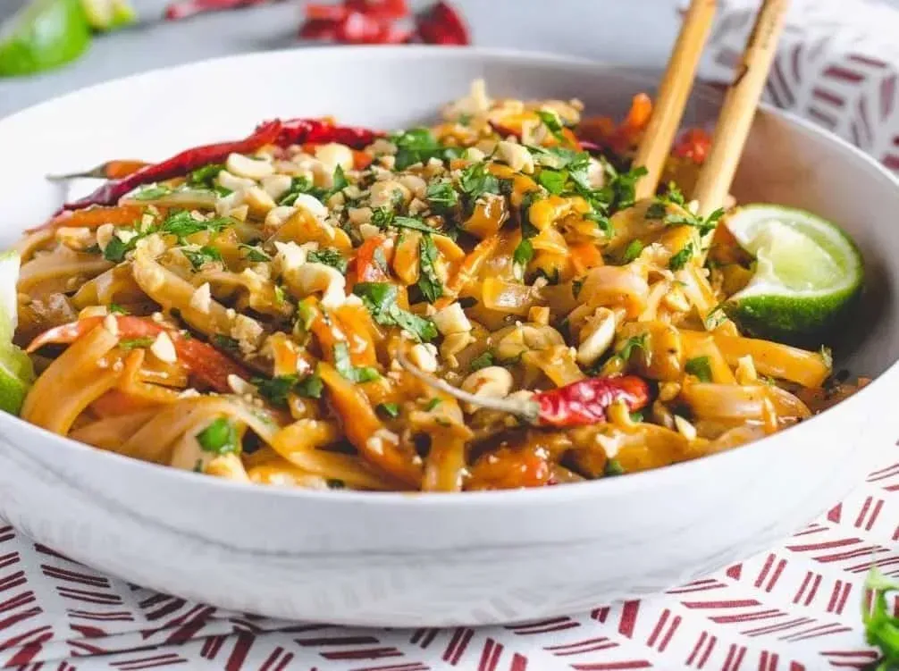 Set on a red-and-white towel, a large earthenware bowl holds the Pad Thai. Two wooden chopsticks are placed in the bowl.