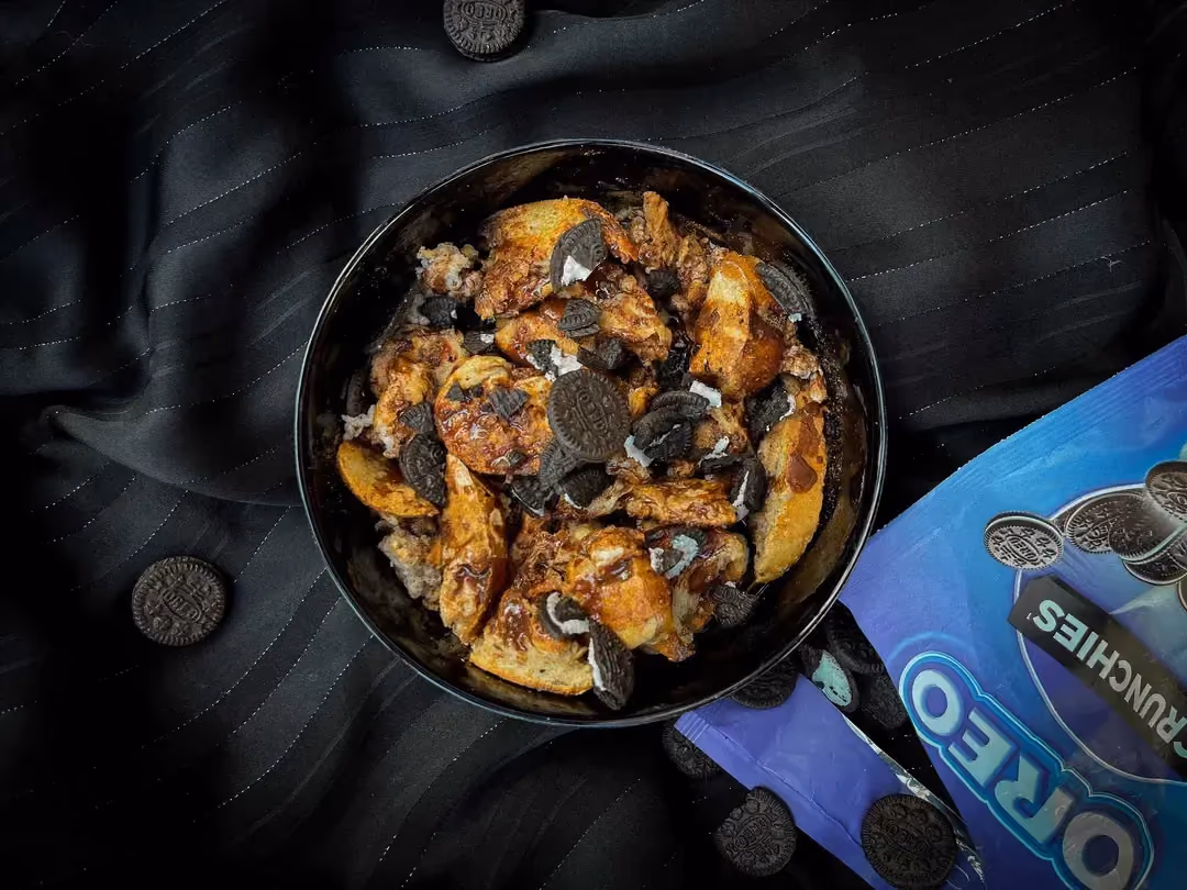 Dans un bol se trouve du pain de mie coupé en petits carrés, de la sauce chocolat et des petits gâteaux Oreo. Un sachet d'Oreo ouvert est à côté.