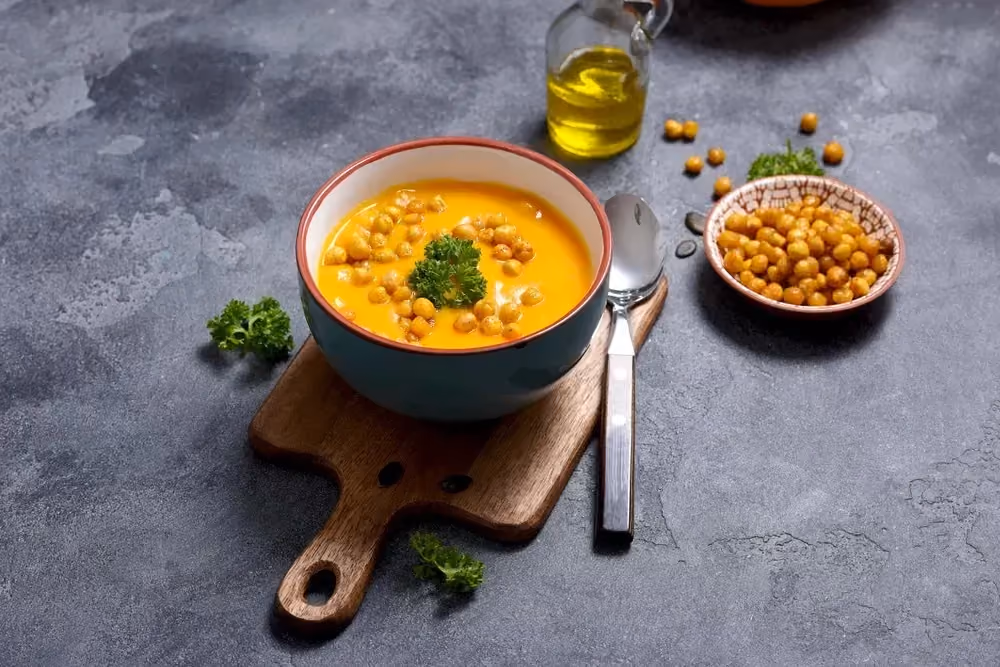 On a wooden cutting board, there is a bowl of chickpea soup. An oil cruet and a small bowl full of chickpeas are next to it. .