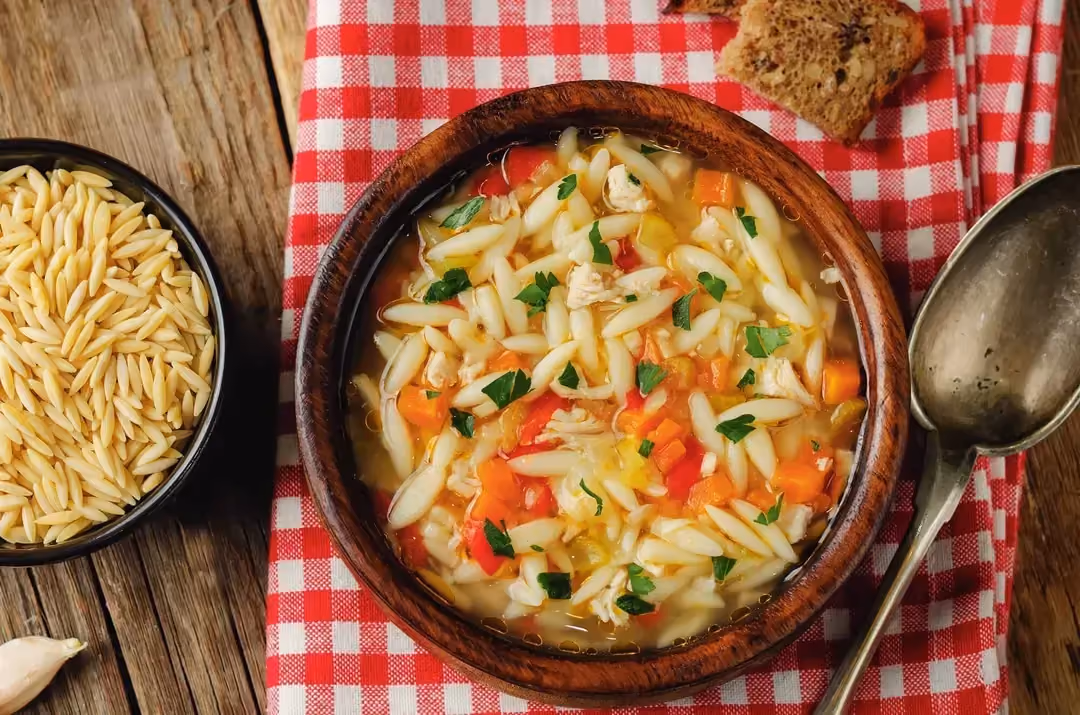 Sur un petit torchon à carreaux rouges et blancs, est posé un gros bol en terre cuite marron contenant la soupe de poulet et les grains d'orzo.