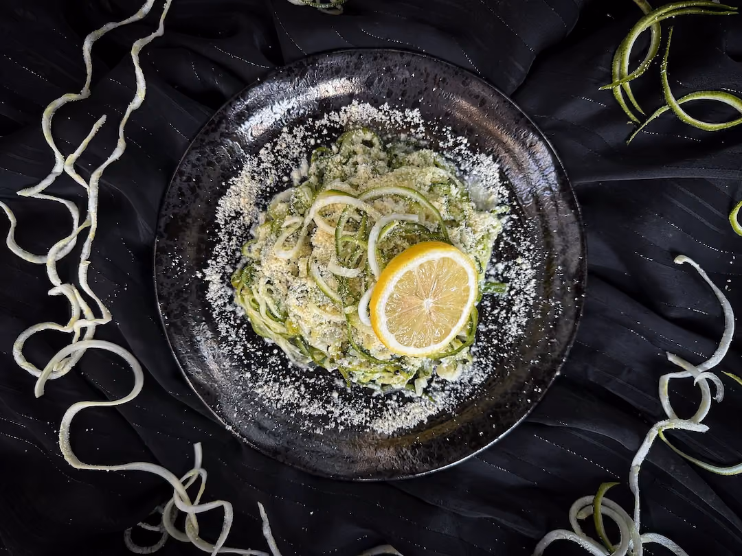 Dans un assiette noire, les courgettes coupées façon spaghettis sont accompagnées d'une tranche de citron jaune, de parmesan et de basilic haché.