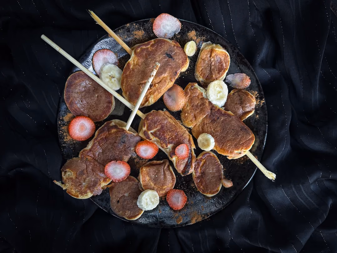 Les sucettes de pancake/banane sont accompagnées de rondelles de fraises et bananes, elles sont déposées sur une jolie assiette noire.