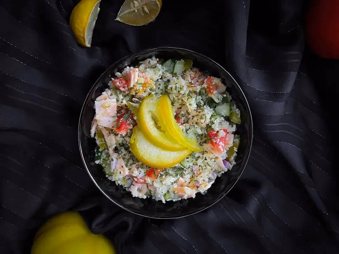 In a small blue bowl are cauliflower, turkey, cucumbers, and tomatoes. Lemon zest is placed on top.