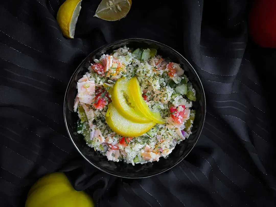 In a small blue bowl are cauliflower, turkey, cucumbers, and tomatoes. Lemon zest is placed on top.