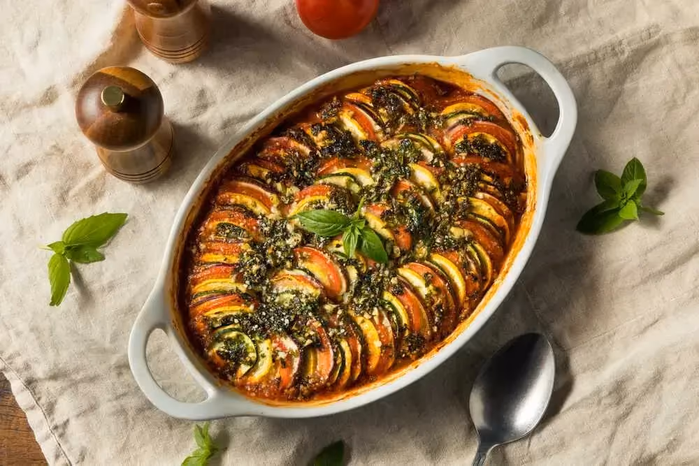 In an oval baking dish, the different vegetables, sliced into rounds, are arranged vertically in alternating layers over a base of tomato sauce.