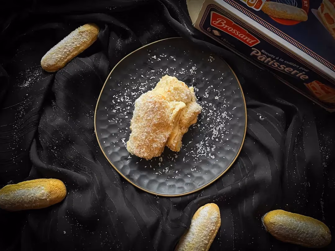 Dans un bol alvéolé de couleur bleu se trouve le tiramisu protéiné coco et citron. Quatre biscuits à la cuillère sont sont à côté du bol.