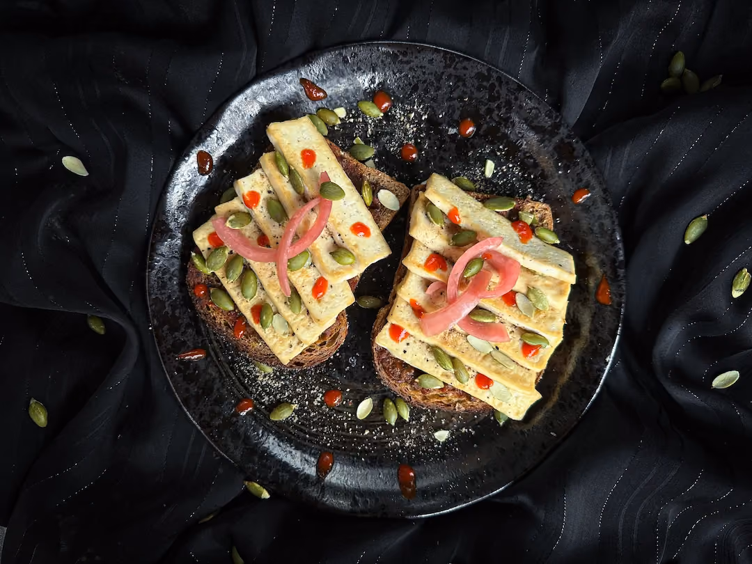In a small bowl, there are small toasts with pesto rosso sauce, tofu, and pumpkin seeds.