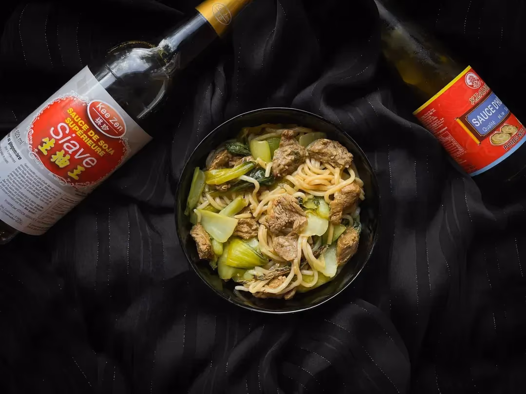 Dans un grand bol se trouve des morceaux de boeuf accompagnés du Pak Choï. Une petite bouteille de Siave et une autre de sauce d'huître sont à côté.