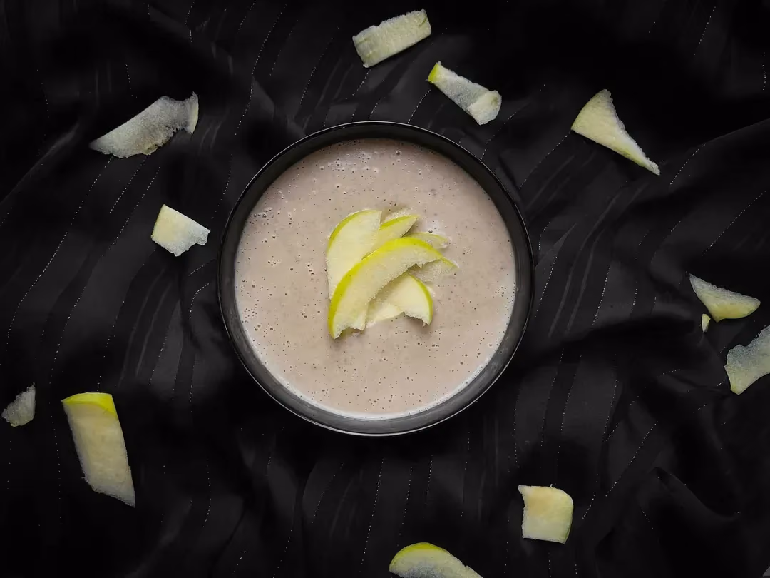 In a bowl is the chestnut velouté, served with thin slices of Granny Smith apple arranged on the soup and beside the bowl.