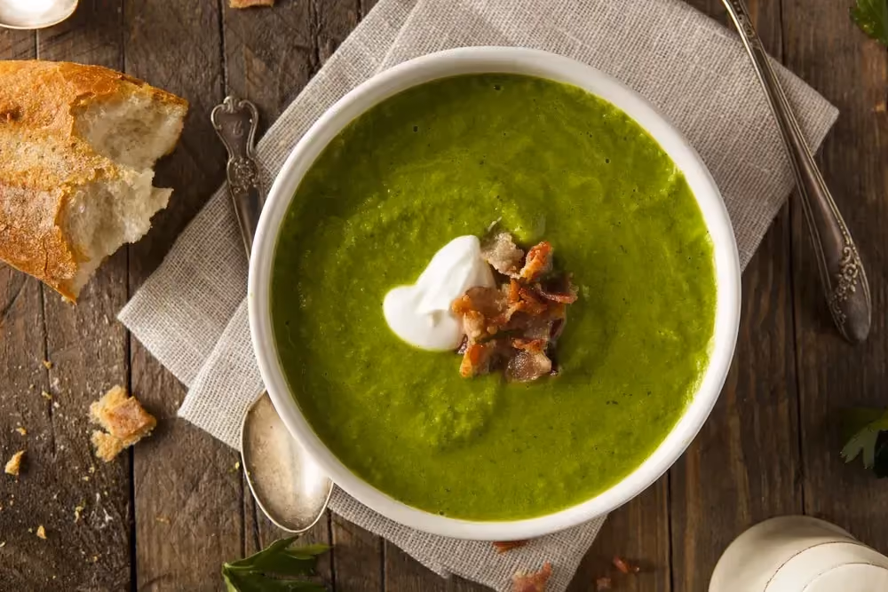 Set on a rustic wooden table is a white bowl filled with pea soup. The green color is especially striking.