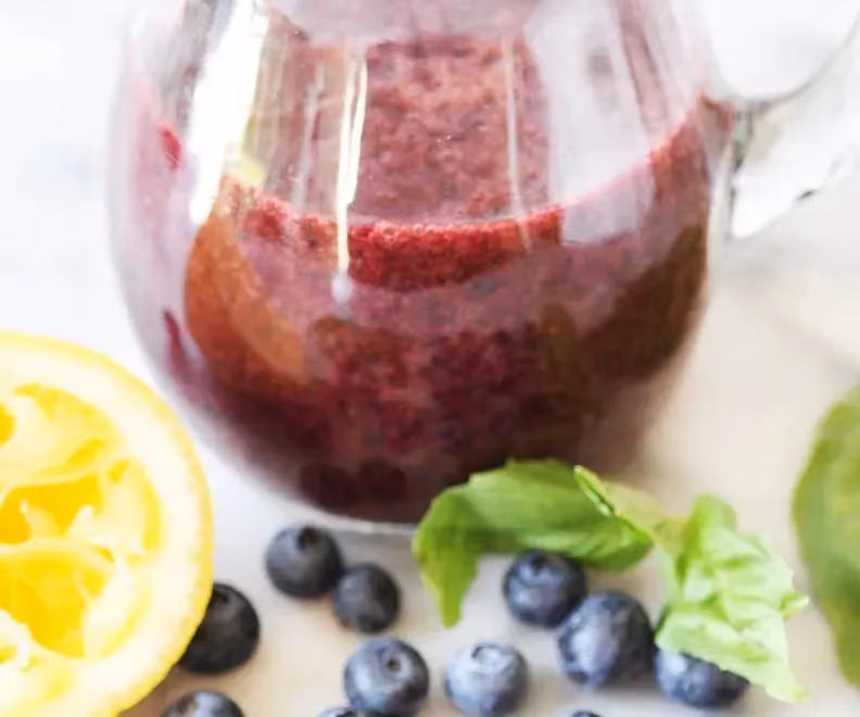 In a large glass jar is the blueberry vinaigrette. Next to it, you can see blueberries, half a yellow lemon, and basil leaves.