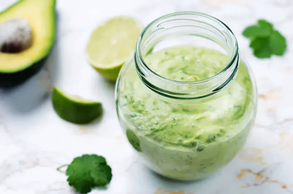 The all-green dressing is in a glass jar that can be sealed. Half an avocado and half a lemon are next to the jar.