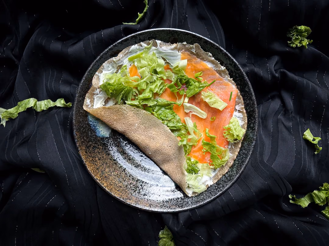 Sur une assiette se trouve une galette de blé noir avec du fromage frais, du saumon fumé et de la ciboulette ciselée. Le wrap n'est pas encore roulé.