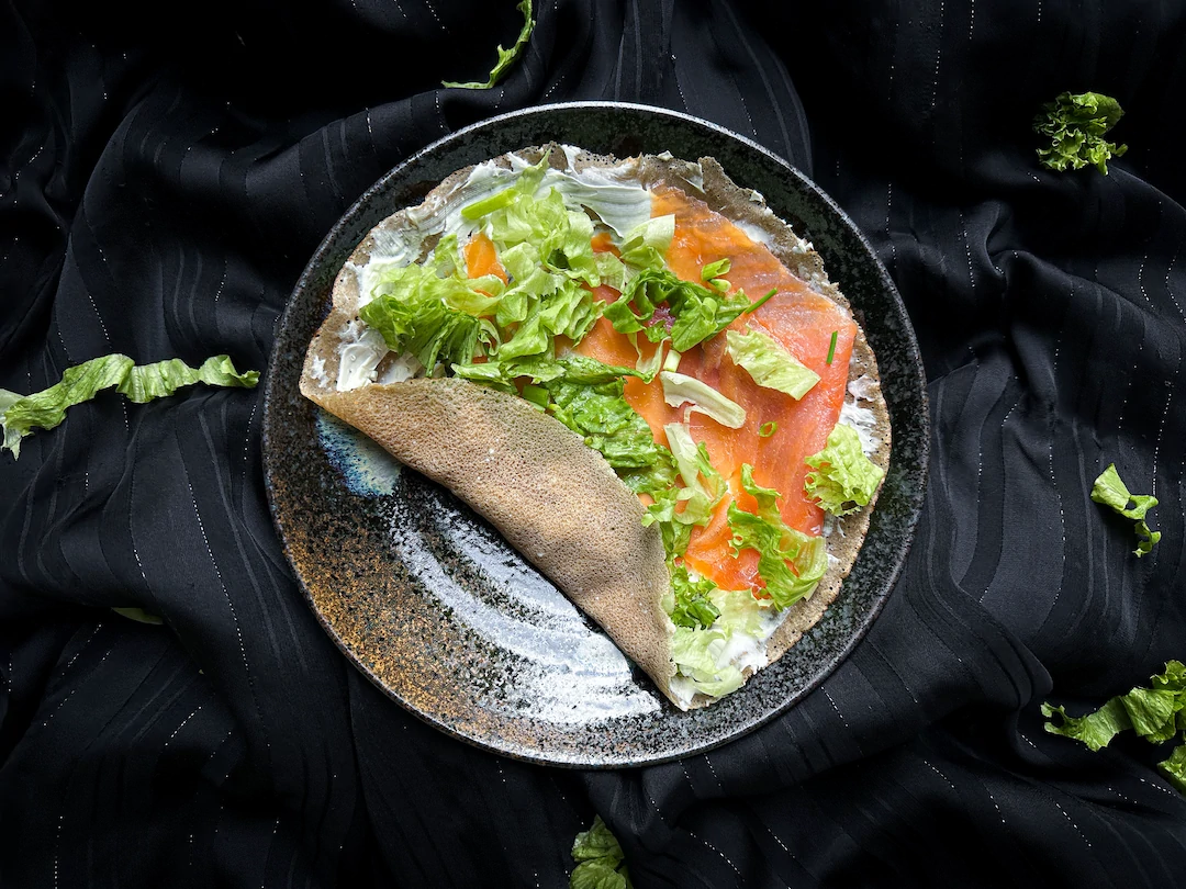 On a plate, there is a buckwheat galette with cream cheese, smoked salmon, and chopped chives. The wrap has not been rolled yet.