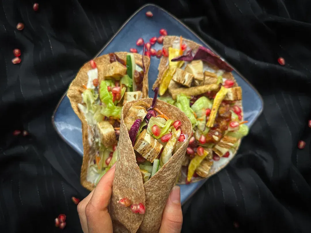 Placed on a black plate are five wraps made with sweet potato, pomegranate, dates, and soy. Around them are half a lime and garlic.