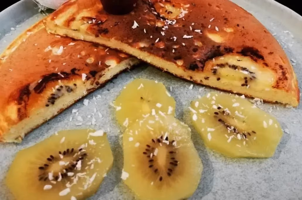 Un Pancake géant coupé en deux laisse apparaître les rondelles de kiwi. A côté se trouve quatre tranches de kiwi avec du coco finement râpé au dessus.