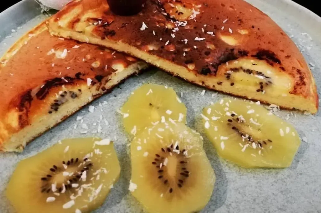 A giant pancake cut in half reveals kiwi slices inside. Next to it are four kiwi slices with finely grated coconut on top.