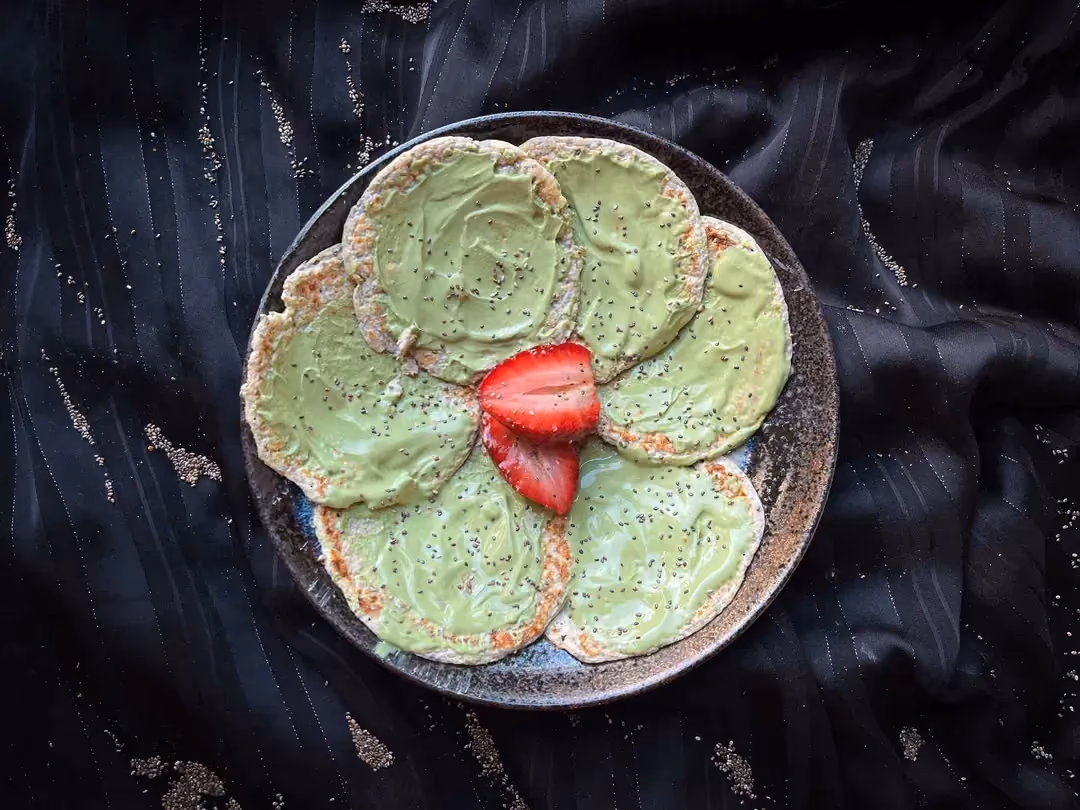 Dans un petit saladier de couleur bleu, se trouve six pancakes aux graines de chia et sauce matcha. Une fraise coupée en deux est au centre du plat.