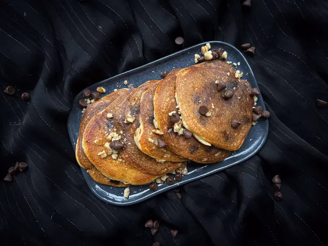 Sur une assiette de couleur noire, les pankakes sont empilés les uns sur les autres. De fines rondelles de bananes sont disposées autour.