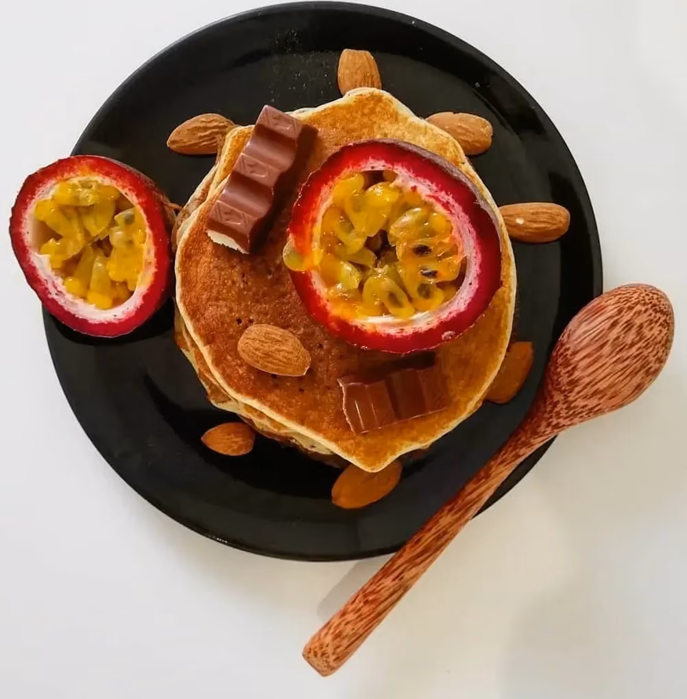 Sur une assiette noire se trouve une pile de pancakes au coco. Un maracuja coupé en deux, une barre de chocolat et des amandes sont visibles.