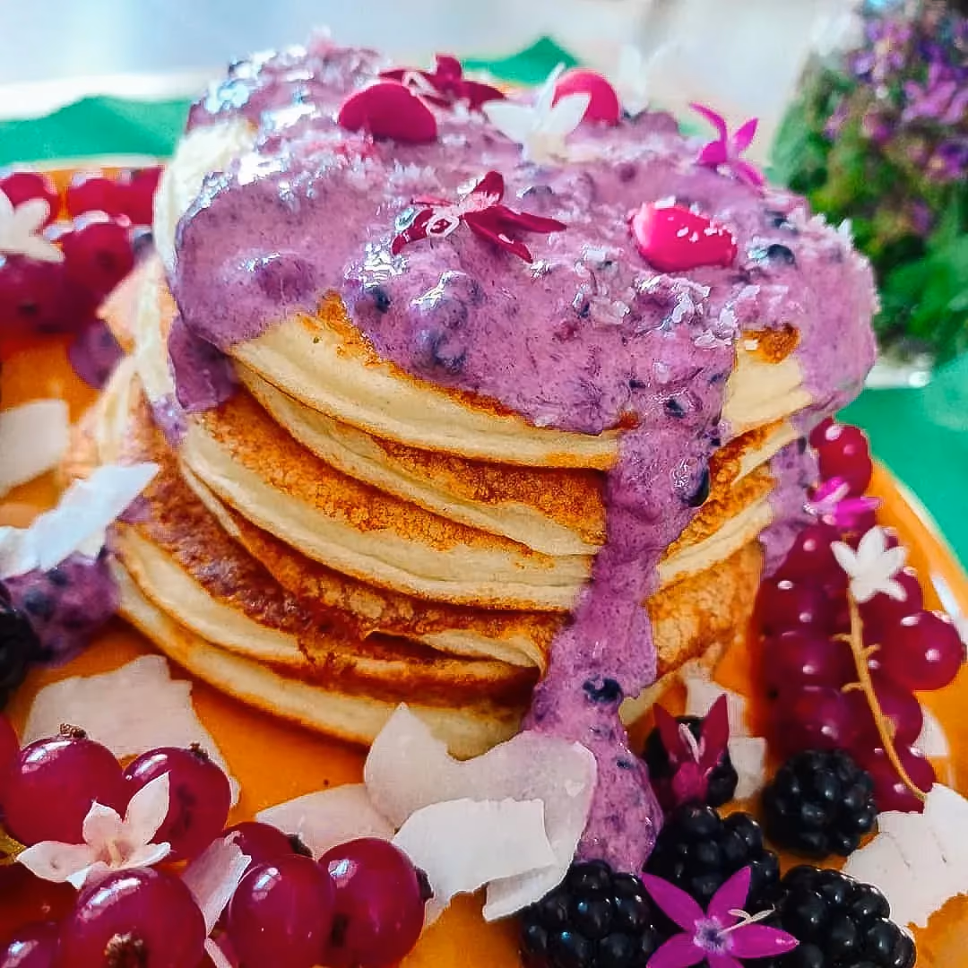The pancakes are stacked on an orange plate and topped with a blackberry topping, with red berries arranged all around as decoration.