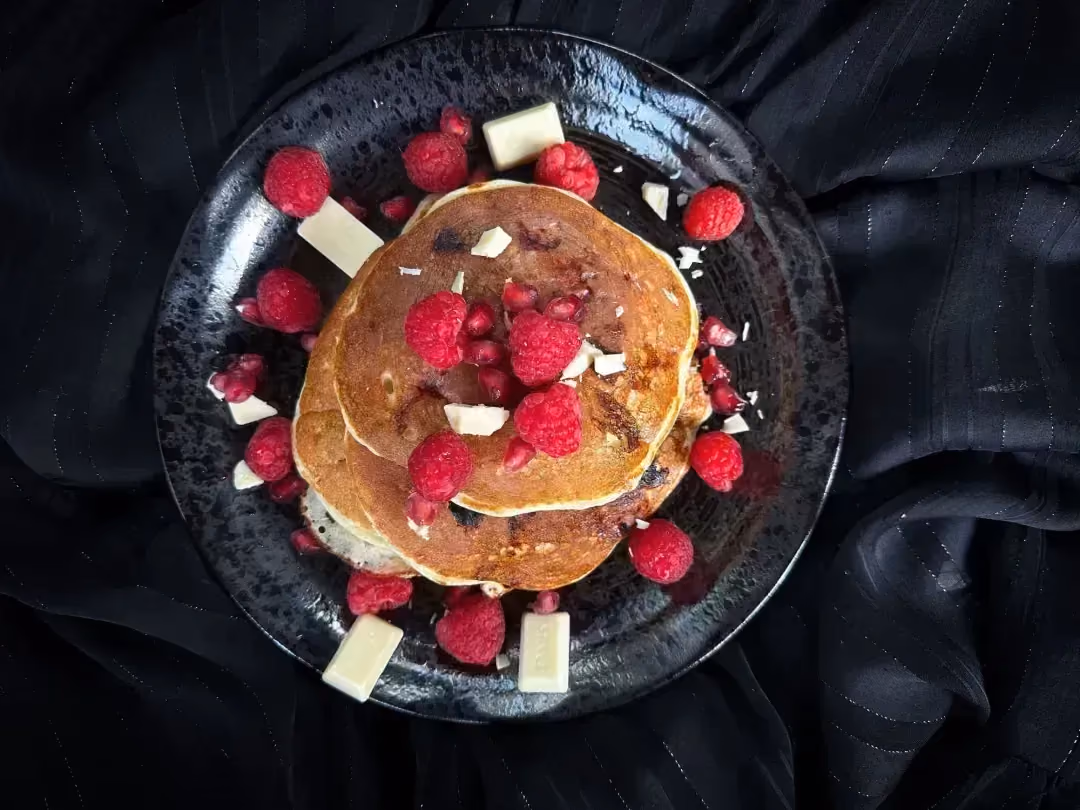 Les pancakes sont empilés au milieux d'un joli petit saladier. les framboises servent de topping et sont dispersées autour des pancakes.