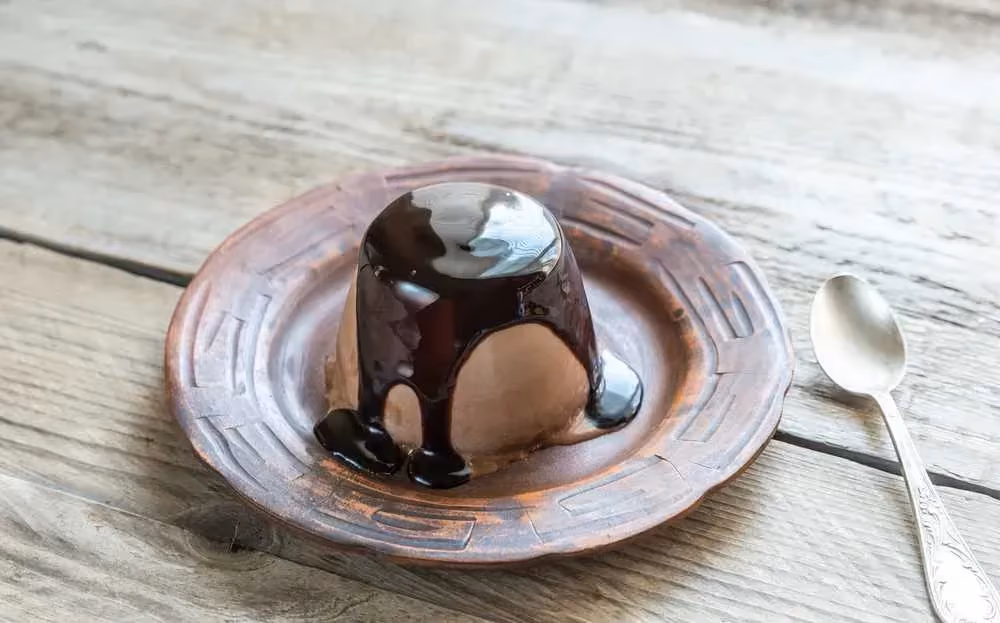 Posée sur une table en bois clair, une assiette creuse de couleur beige accueille la Panna cotta recouverte par un chocolat coulant.