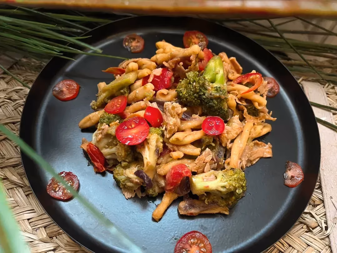 On a shallow plate, there is broccoli pasta with bacon, chicken, and cherry tomatoes.