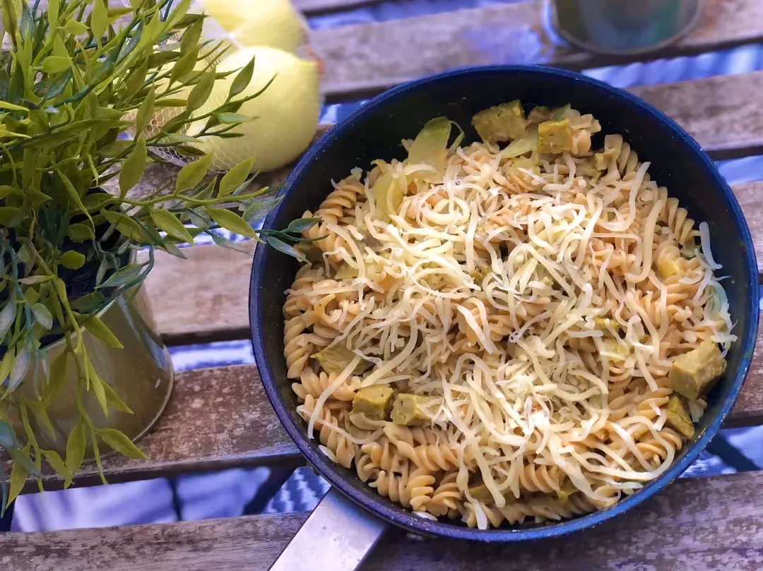 Set on a garden table, you can see a pan with red lentil pasta covering the seitan. Two lemons are placed next to it.