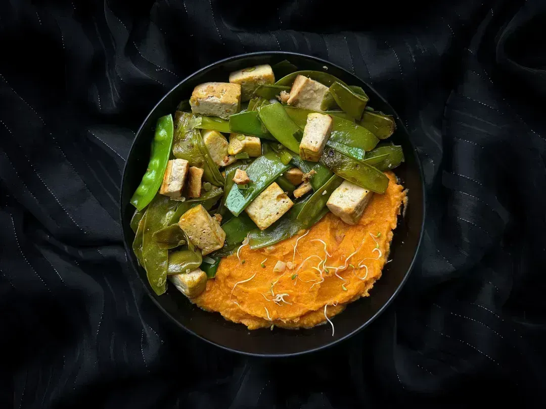 This very colorful recipe, a tofu, flat green bean, and sweet potato skillet, is presented in a small blue bowl.
