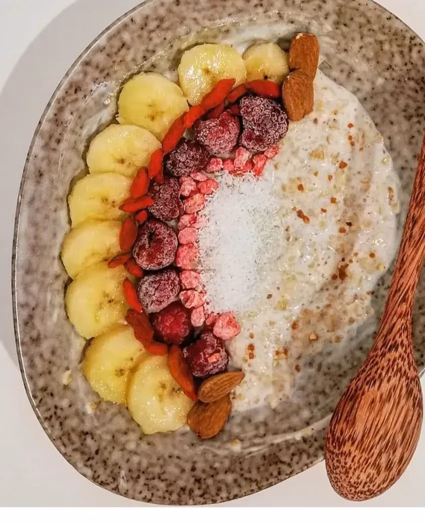 In an oval beige earthenware dish, the porridge is half covered with thin banana slices, a few berries, and almonds.