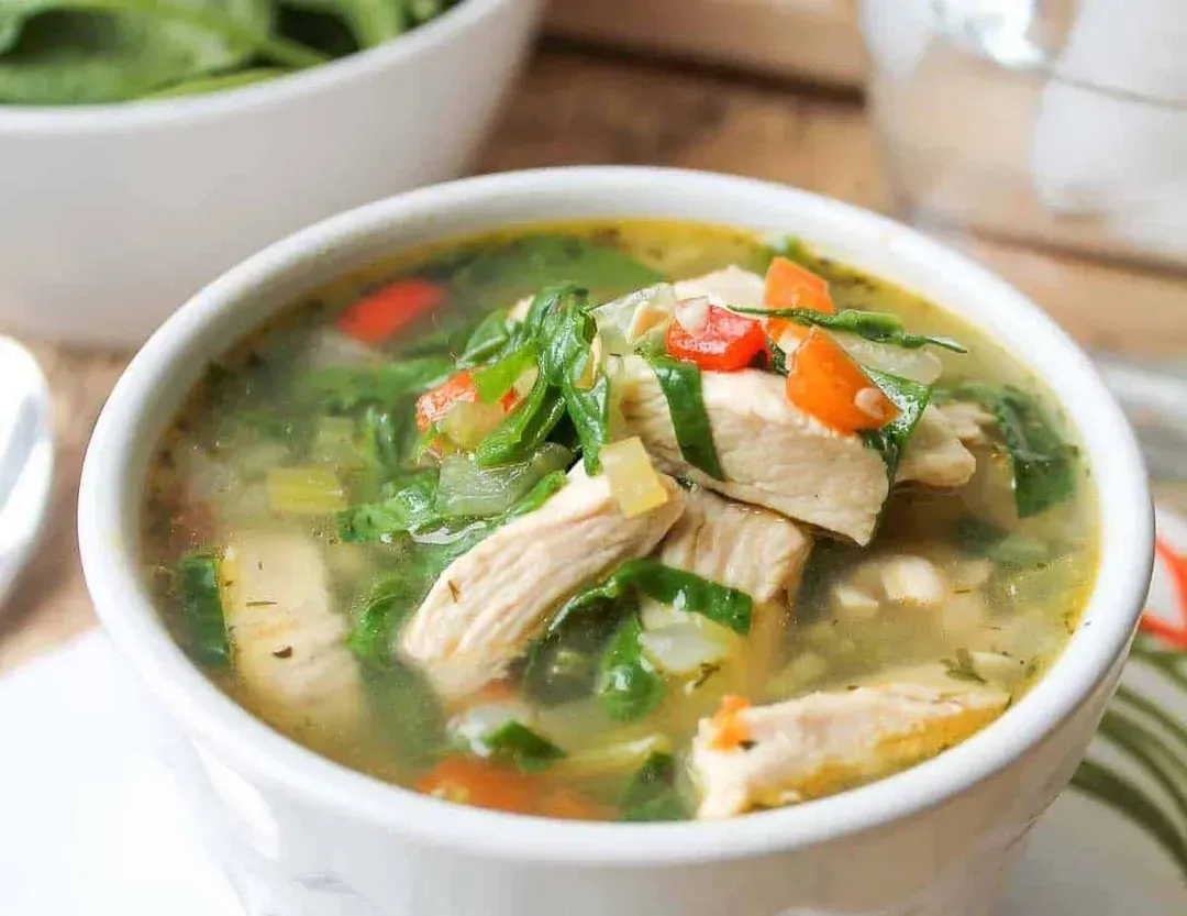 Large pieces of chicken accompanied by spinach make up this chicken soup, presented in a large, wide white earthenware bowl.