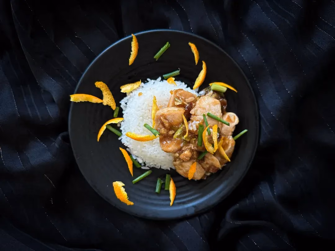 A terracotta bowl sits on a rustic wooden table. It contains white rice on one side and well-grilled chicken on the other.