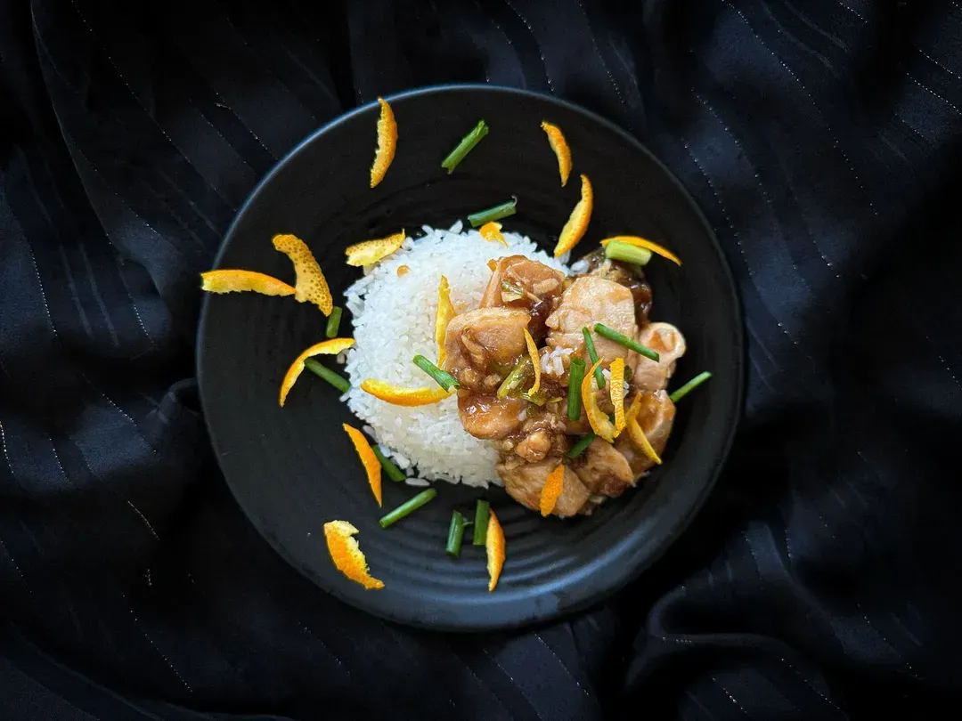 A terracotta bowl sits on a rustic wooden table. It contains white rice on one side and well-grilled chicken on the other.