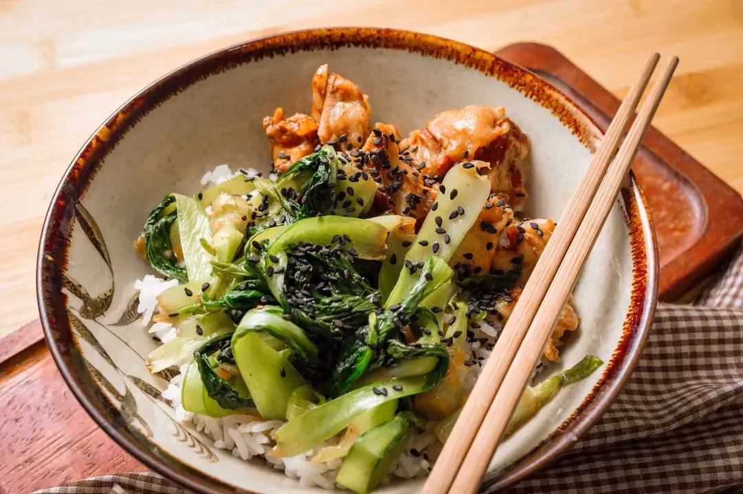 Teriyaki chicken and pak choi sit on top of a ball of rice, all served in a large bowl with two chopsticks.