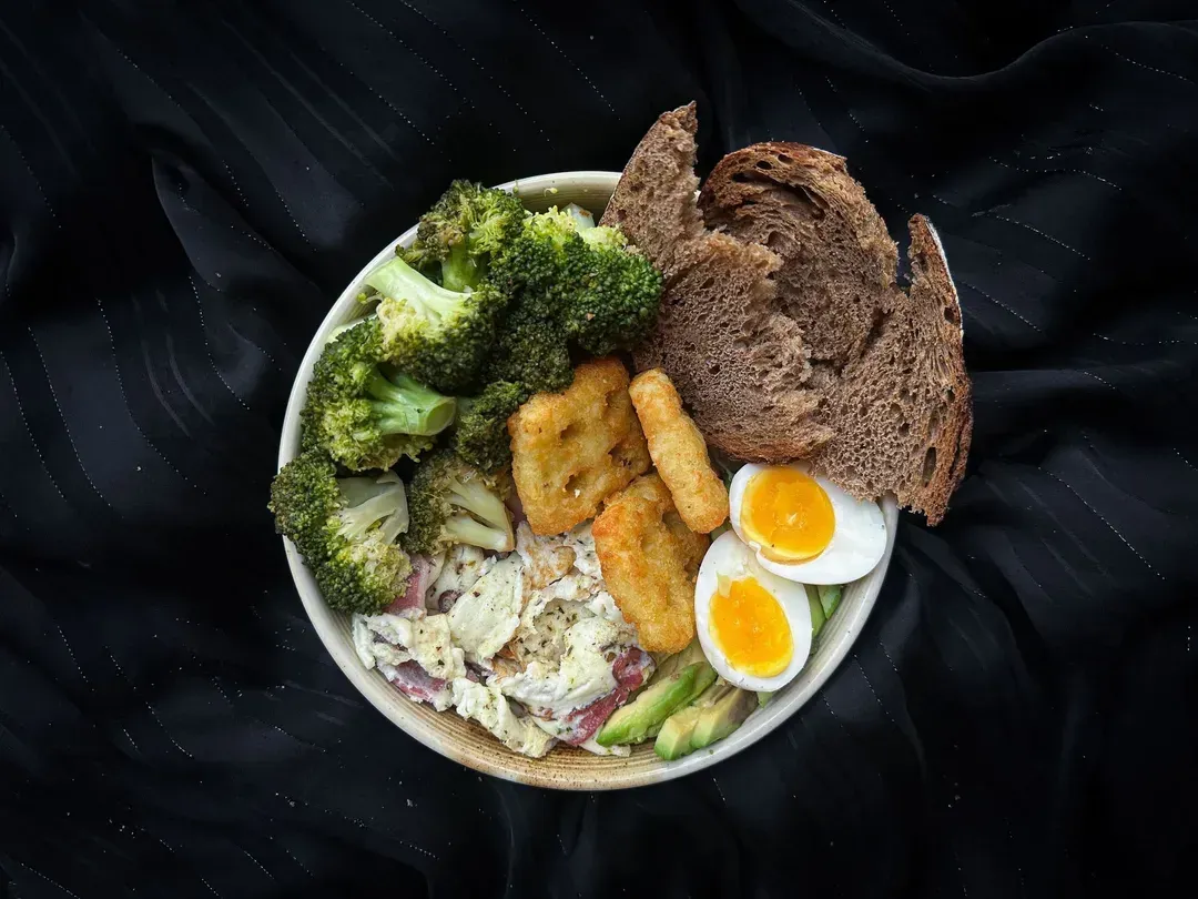 A plate contains the Power Bowl made with eggs, bacon, broccoli, avocado, as well as two rösti patties and a slice of rye bread.