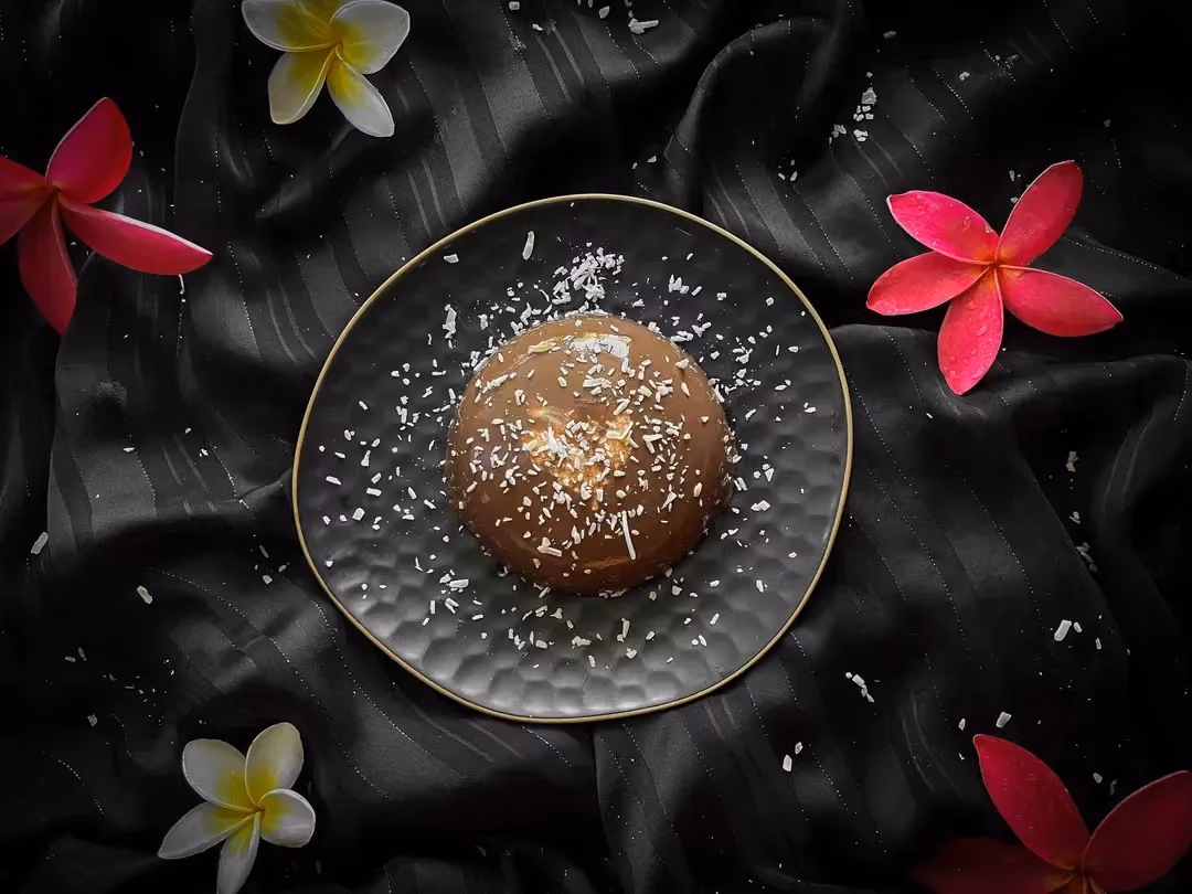 Le pudding choco et coeur surprise coco est présenté dans un saladier de couleur bleu alvéolé. Quelques fleurs de frangipaniers sont autour du plat.