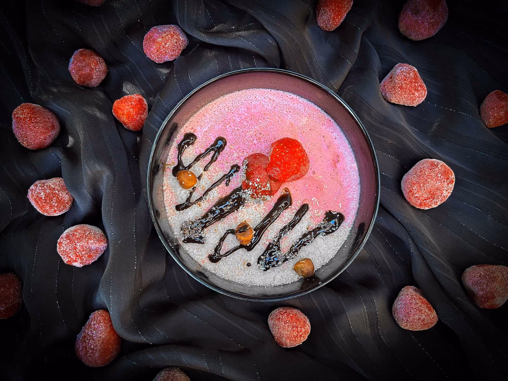 In a bowl is the strawberry and skyr pudding with its beautiful pink color. The topping is made of strawberries and a drizzle of dark chocolate sauce.