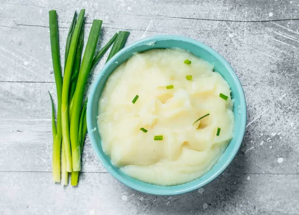 Placed on a wooden table, a small green bowl contains the celery purée. A few peeled celeries are placed next to the bowl.