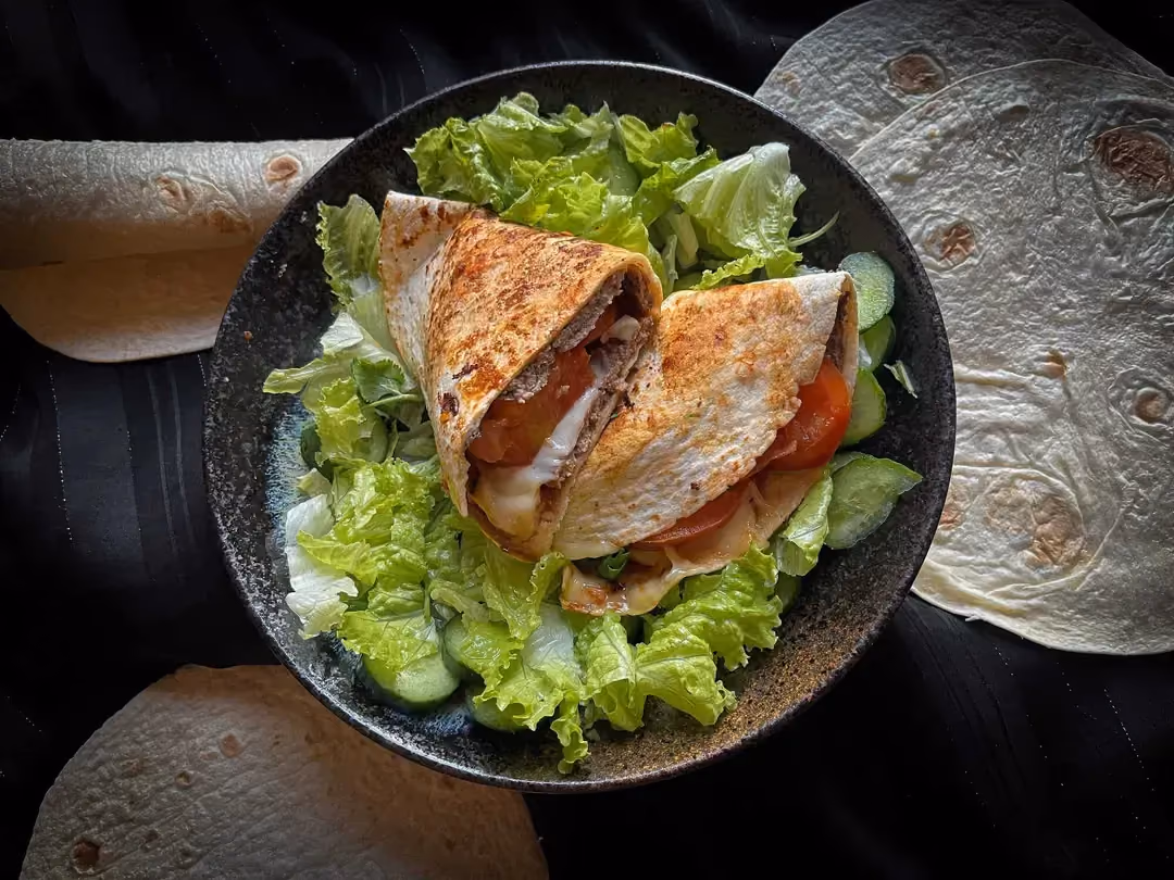 In a small bowl, on a bed of iceberg lettuce, there are two tortillas filled with beef, cherry tomatoes, and Emmental cheese