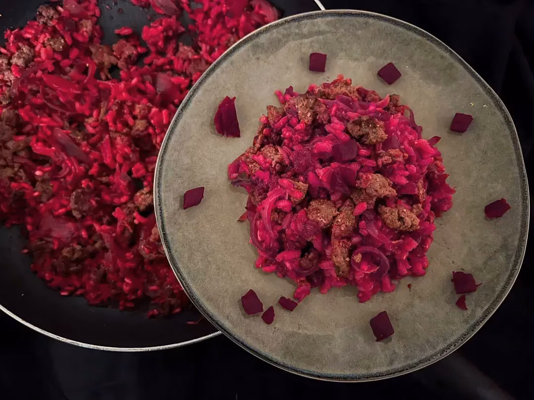Une assiette blanche est prête à être servie, elle se trouve à côté de la poêle qui contient le risotto à la betterave et la viande de boeuf hachée.