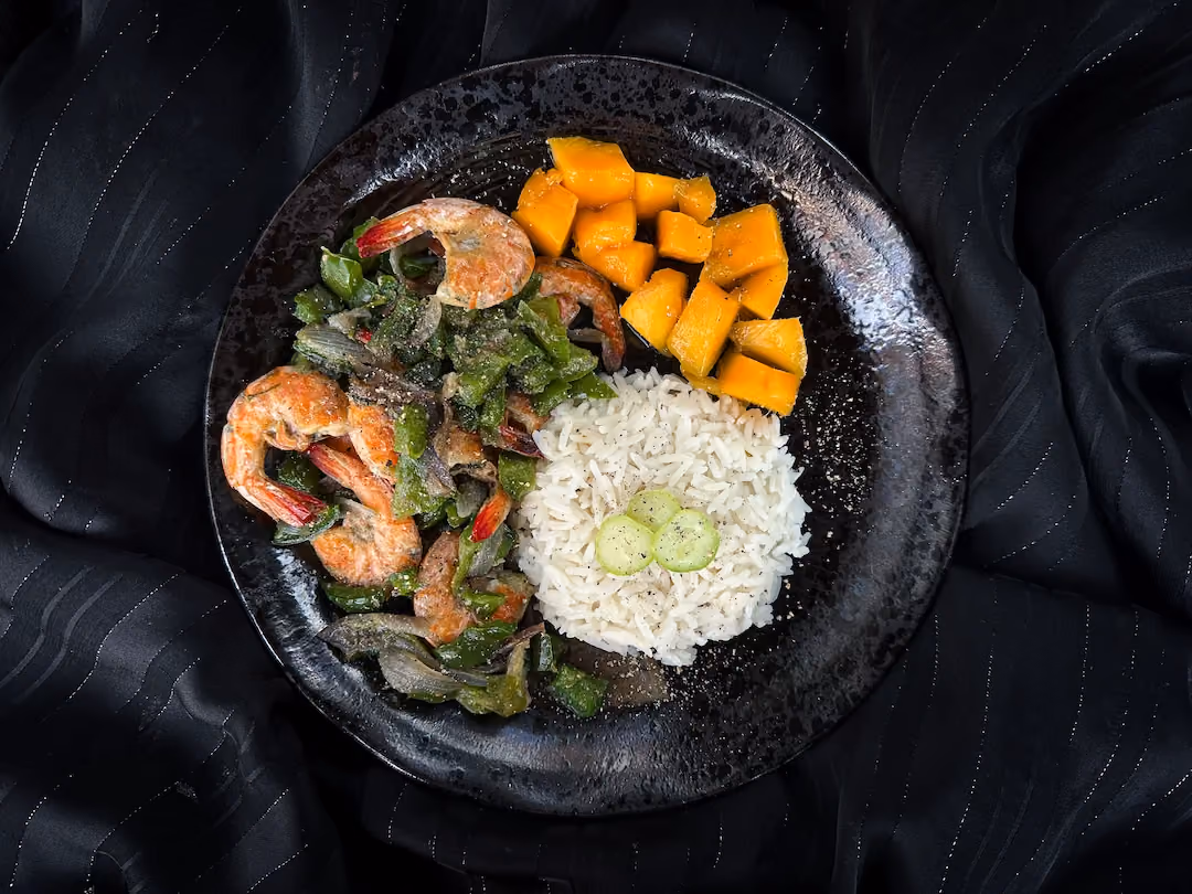 Dans une assiette noire, se trouve des crevettes et poivrons, des mangues coupées en cube accompagnés de lait de coco.